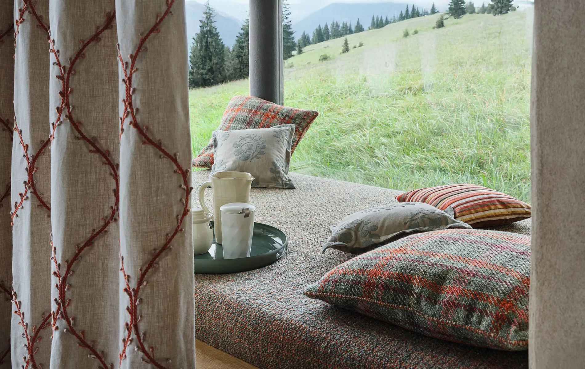 Leinenvorhang mit gesticktem Rankenmuster vor einer Fensterbank mit Kissen und Bergblick